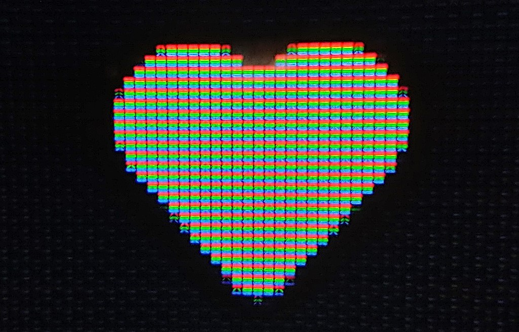 A very close up photo of a small section of a screen with a white heart icon. You can make out that the screen is a grid and the grid squares where the heart is have red, green, and blue bands, which look white when seen from a distance